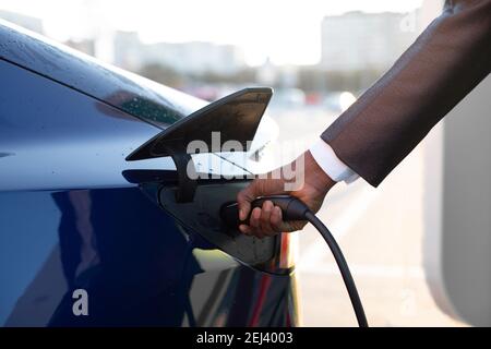 Nahaufnahme der Hand des Menschen, das Einführen eines Ladegeräts in sein Elektroauto vor dem Hintergrund verschwommener Stadtgebäude Stockfoto