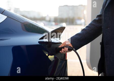 Nahaufnahme der Hand des Menschen, das Einführen eines Ladegeräts in sein Elektroauto vor dem Hintergrund verschwommener Stadtgebäude Stockfoto