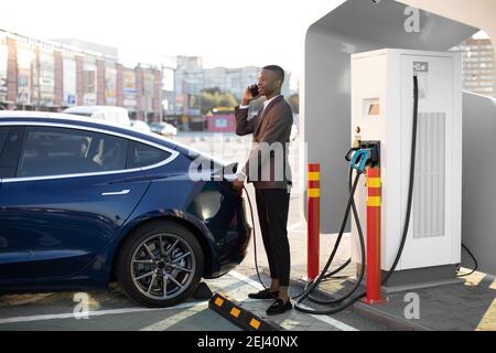 Seitenansicht des jungen hübschen afrikanischen Mannes im Business-Anzug, der Kabel in die Autosteckdose einsteckt, um sein neues modernes Luxus-Elektroauto im Freien aufzuladen Stockfoto