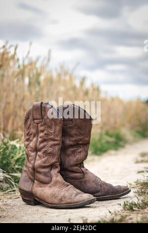 Cowboy Boot auf Feldweg neben Weizenfeld. Alte abgenutzte Lederschuhe im Freien. Retro-Stil noch Leben der Schuhe in ländlichen Szene mit launischen Himmel Stockfoto