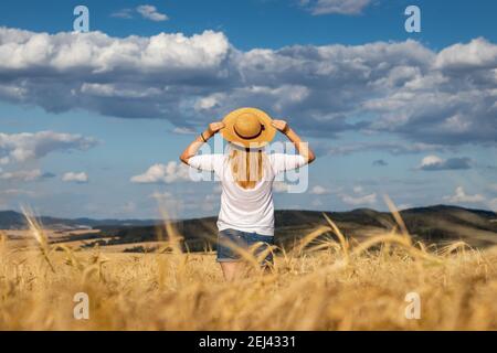 Glückliche Frau mit Strohhut im Weizenfeld stehen und genießen den Sommer. Blonde Haare Frau Gefühl Freiheit. Wunderschöner Himmel. Stockfoto