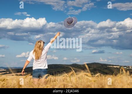 Happy blonde Haare Frau werfen blauen Sonnenhut gegen schönen Himmel. Genießen Sie den Sommer. Sorglose Frau, die im Weizenfeld steht. Stockfoto