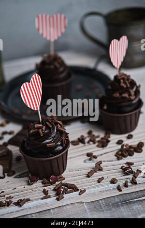 Mehrere Muffins oder Cupcakes mit schokoladenförmiger Sahne am weißen Tisch. Eine Karte in Form eines Herzens zum Valentinstag. Die Hand einer Frau zerbröckelt Stockfoto