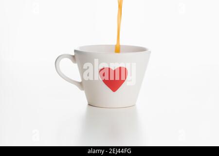 Kaffee wird von oben in einen weißen Becher mit einem roten Liebesherz gegossen, der auf einer schlichten weißen Oberfläche und einem weißen Hintergrund isoliert ist. Stockfoto