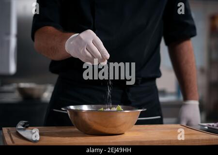 Koch kocht Hände in Handschuhen und fügt Salz in griechischen Salat hinzu Im Restaurant Küche Stockfoto