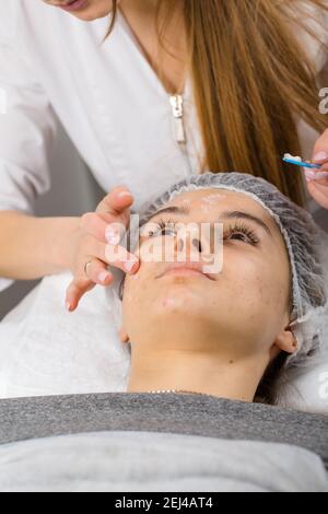 Kosmetikerin oder Dermatologin, die Creme mit der Hand auf das Gesicht während der Gesichtsmassage für eine entspannte junge Frau im Schönheitssalon, Rückansicht. Stockfoto