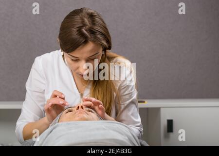 Kosmetikerin oder Dermatologin, die Creme mit der Hand auf das Gesicht während der Gesichtsmassage für eine entspannte junge Frau im Schönheitssalon, Rückansicht. Stockfoto