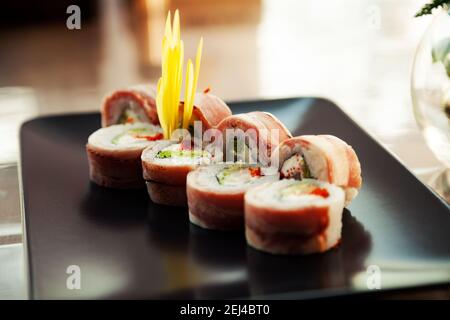 Sushi-Rolle mit Speck und gebratenem Seebarsch auf einem schwarzen Teller, Zutaten gebratener Seebarsch, Frischkäse, Speck, Iceberg-Salat, fliegender Fischrogen, Teriyaki Sauce, Reis, Nori. Für die Speisekarte des Restaurants Stockfoto