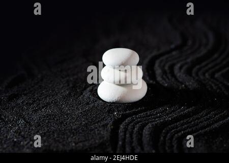 Steingarten, weiße Kieselsteine auf schwarzem Sand, Wellenmuster. Pyramide aus drei glatten weißen Steinen, Meereskiesel und vulkanischem Sand, schwarzer Hintergrund. Stockfoto