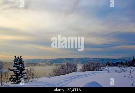 Blick auf ein schneebedecktes Flussufer und bewaldete Berge in der Dämmerung. Stockfoto