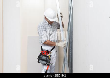 Man construction worker or plasterer holding drywall metal profiles near plasterboard white wall in building site. Wearing white hardhat, work gloves, Stockfoto