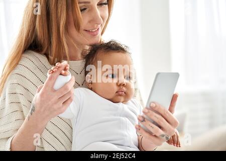 Glücklich junge kaukasische Mutter hält niedlich afrikanische Baby Tochter mit Telefon. Stockfoto