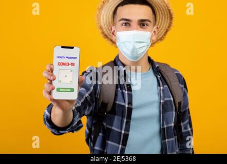 Männlicher Tourist in Gesichtsmaske zeigt online covid-19 Immunität Pass auf Smartphone, oranger Hintergrund, selektiver Fokus Stockfoto
