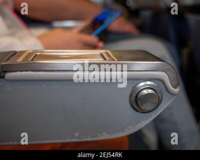 Nahaufnahme eines runden Metallknopfes auf einer Armstütze, um zurückzufallen und sich an einem Flugzeug zu erholen. Person, die auf einem Passanten-Sitz sitzt und einen Smarth hält Stockfoto