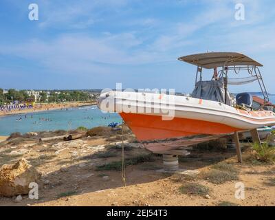 Oranges und weißes Motorboot, das auf Reifen und Holzstützen am Ufer in Ayia Napa steht. Mittelmeer im Hintergrund. Stockfoto