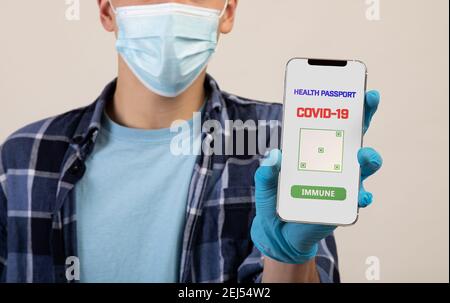 Unerkennbarer junger Mann in Gesichtsmaske mit Handy mit covid-19 Gesundheitspass auf hellem Studiohintergrund Stockfoto