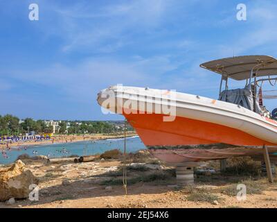 Oranges und weißes Motorboot, das auf Reifen und Holzstützen am Ufer in Ayia Napa steht. Mittelmeer im Hintergrund. Stockfoto