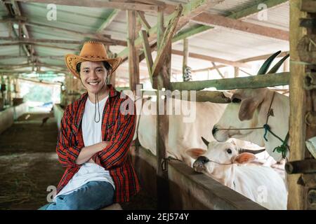 Lächelnder Mann mit Cowboyhut sitzt und schaut auf die Kamera mit gekreuzten Händen im Hintergrund der Kuh Farmstall Stockfoto