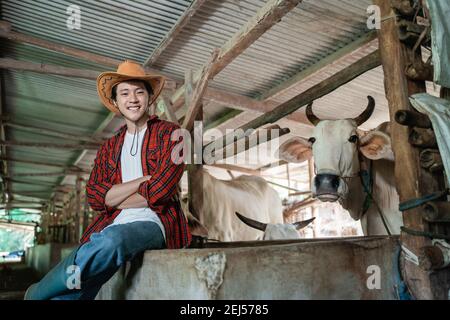 Lächelnder Cowboymann, der einen Cowboyhut trägt, während er in der Nähe sitzt Die Kuh Schuppen mit gekreuzten Händen im Hintergrund Der Kuhstall Stockfoto