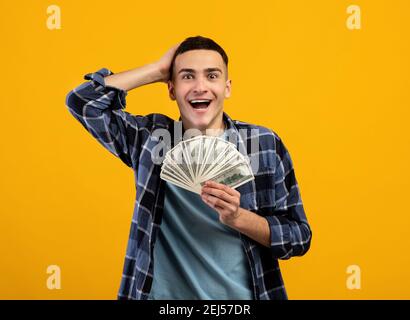 Aufgeregt junger Mann genießen großen Jackpot, hält viel Geld, kann nicht glauben, sein Glück auf orangen Studio Hintergrund Stockfoto