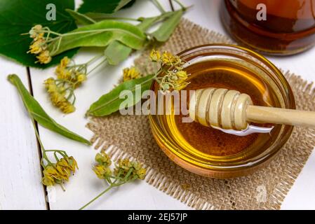 Stick für Honig in Glasschale mit Lindenhonig und frischen Lindenblüten auf einem Holztisch. Selektiver Fokus. Stockfoto