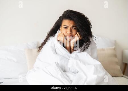 Enttäuschte Latino-Frau mit dunkler Lockenfrisur im Bett sitzend, faltete ihre Stirn, legte den Kopf auf ihre Hände, schaute geradeaus und wickelte Stockfoto