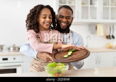 Fröhlicher afrikanischer Vater lehrt Tochter, wie man Salat zubereitet Stockfoto