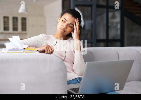 Müde multirassische junge Frau, die auf dem Sofa mit Laptop sitzt und wegschaut, faltige Stirn, Hand mit dem Kopf haltend, überarbeitet, hält Stockfoto