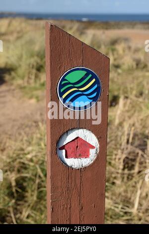 Isolierter Holzpfosten mit Fife Coastal Path-Logo und Pfeil mit verschwommenem Hintergrund aus Seegras, Sand, Meer und blauem Himmel Stockfoto