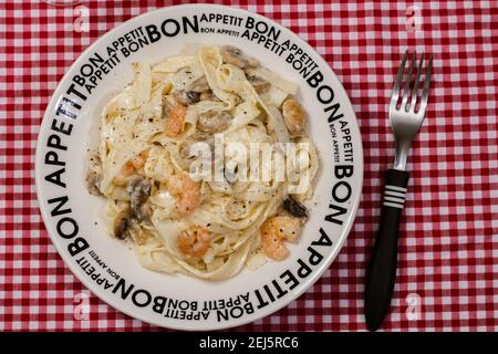 Köstliche Fettuccine mit Sahne Sauce von Pilzen und Garnelen auf einem Teller, der Bon Appetit mit rot karierten Tischdecke sagt. Italienische Küche. Stockfoto