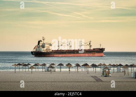 Setúbal, Portugal - 29. August 2020: Frachtschiff am Figueirinha Strand in Setúbal, Portugal, bei Sonnenuntergang, mit den Sonnenschirmen und dem Sand im f Stockfoto