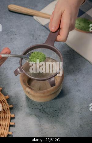 Vertikales Bild Matcha-Tee-Pulver mit Sieb in einer Holzschale auf Graustein Hintergrund streuen. Japanischer Grüner Tee. Zubereitung Von Matcha-Tee 2021. Stockfoto