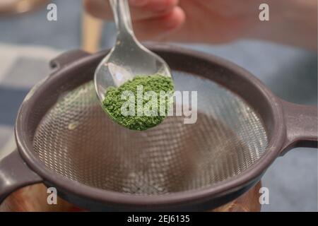 Vertikales Bild Matcha-Tee-Pulver mit Sieb in einer Holzschale auf Graustein Hintergrund streuen. Japanischer Grüner Tee. Zubereitung Von Matcha-Tee 2021. Stockfoto