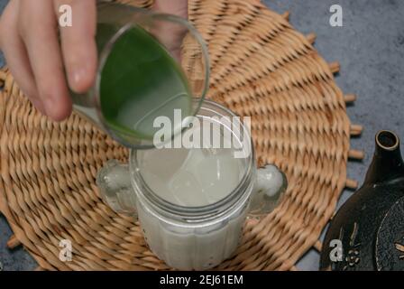 Horizontales Bild von Hand gießt grünen Tee über Eismilch auf grauen Stein Hintergrund und Teekanne. Japanischer Grüner Tee Konzept. Zubereitung Von Matcha-Tee 2021. Stockfoto