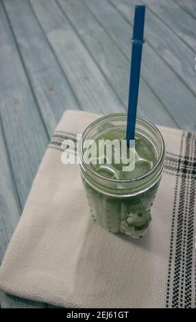 Schließen Sie das vertikale Bild von Kristallglas voller Matcha-Tee, gemischt mit Zucker und Honig auf blauem Holzhintergrund mit einem Kaminsims. Zubereitung von Matcha-Tee. Stockfoto