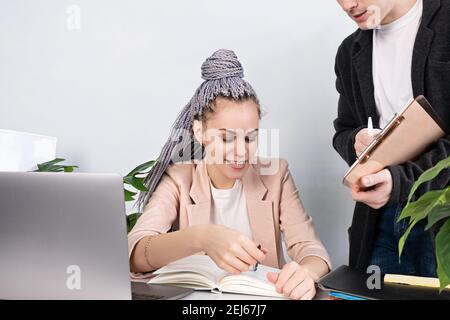 Junge selbstbewusste Frau Führer erklärt etwas zu ihrem Assistenten, zeigt in einem Notebook, während im Büro in einer Jacke auf einen Arbeitstisch und l sitzen Stockfoto