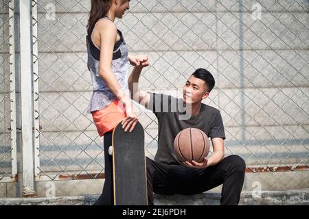 Junge asiatische Erwachsene männliche Basketballspieler und weibliche Skateboarder stoßen Fäuste auf dem Platz im Freien Stockfoto