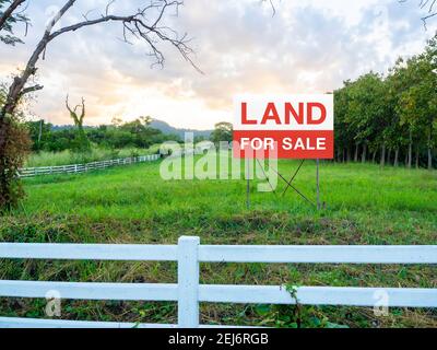 Land zum Verkauf Zeichen auf leerem Land, grüne Wiese in der Nähe des Baumes und umgeben mit weißem Holzzaun auf schönen Himmel Hintergrund. Rote und weiße Buchstaben Stockfoto