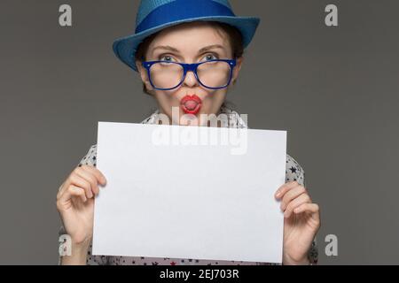 Schöne Brünette Mädchen mit Brille hält ein weißes Blatt in den Händen und ist auf einem grauen Hintergrund überrascht Stockfoto