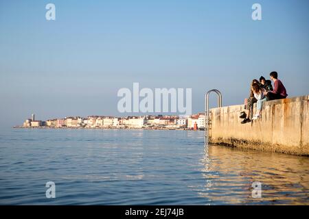 Gruppe von jungen Menschen hängen an einem Betonsteg, malerische Altstadt Piran an der slowenischen adriaküste im Hintergrund, Slowenien, Europa Stockfoto