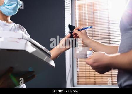 Der Mann signiert eine Unterschrift auf einem Smartphone, um ein Paket von einem Anbieter zu erhalten. Männlicher Kurier mit einer Maske, die einen Karton liefert. Das Konzept schnell Stockfoto