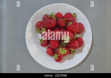 Ein Bündel von reifen, reifen rot-kastanienbraunen Erdbeeren, die auf einer weißen Keramikplatte auf einer grau-silbernen Korbtischdecke liegen, Stockfoto