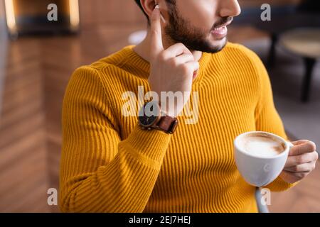 Beschnittene Ansicht des arabischen Mannes, der den Kopfhörer anpasst, während er die Tasse hält Kaffee im Restaurant Stockfoto