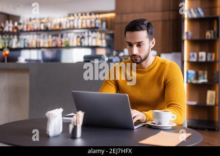 Fokussierter arabischer Freiberufler, der an einem Laptop in der Nähe einer Tasse Kaffee im Restaurant arbeitet, verschwommener Vordergrund Stockfoto