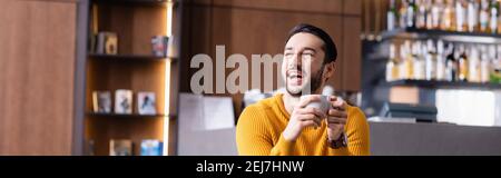 Aufgeregter araber, der lachte, während er eine Tasse Kaffee hielt und im Restaurant wegschaute, Banner Stockfoto