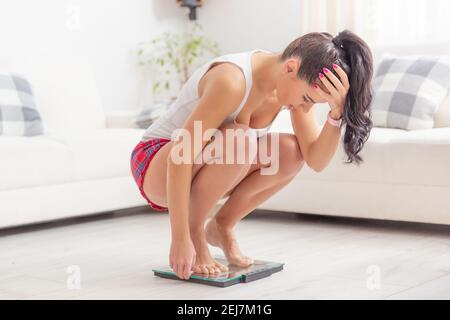 Junge Frau hockt auf einer Waage, die ihren Kopf über Gewichtszunahme frsutriert hält. Stockfoto