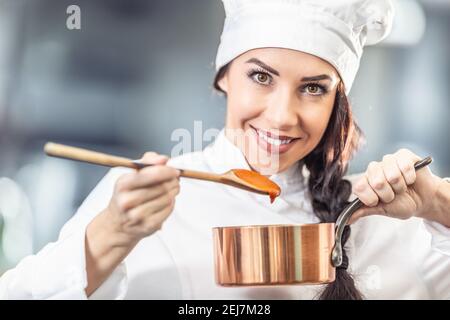 Die professionelle Köchin hält einen Holzlöffel mit Orangensauce und eine Pfanne mit Hut und Chefuniform. Stockfoto