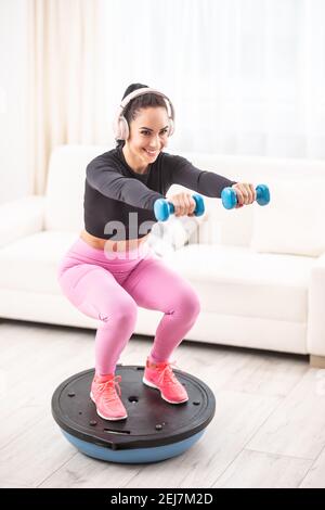 Passen schöne Frau in ein Trainingsoutfit und Kopfhörer hockt zu Hause auf einem Balance Ball halten Hanteln. Stockfoto