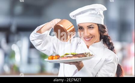 Frau in Chefuniform gießt Sauce über Steak mit Gemüse in der Küche. Stockfoto