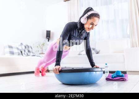 Passform eine gut aussehende Frau trägt Kopfhörer während ihres Heimtrainings und hält die Plank mit den Händen auf einem Gleichgewichtssball. Stockfoto